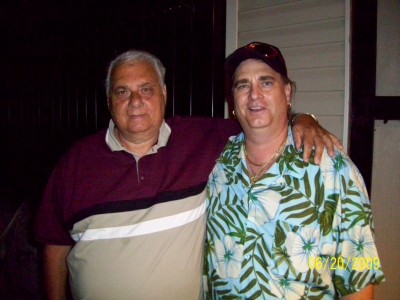 Two men stand side by side outdoors at night, with the younger man's arm around the older man's shoulder.